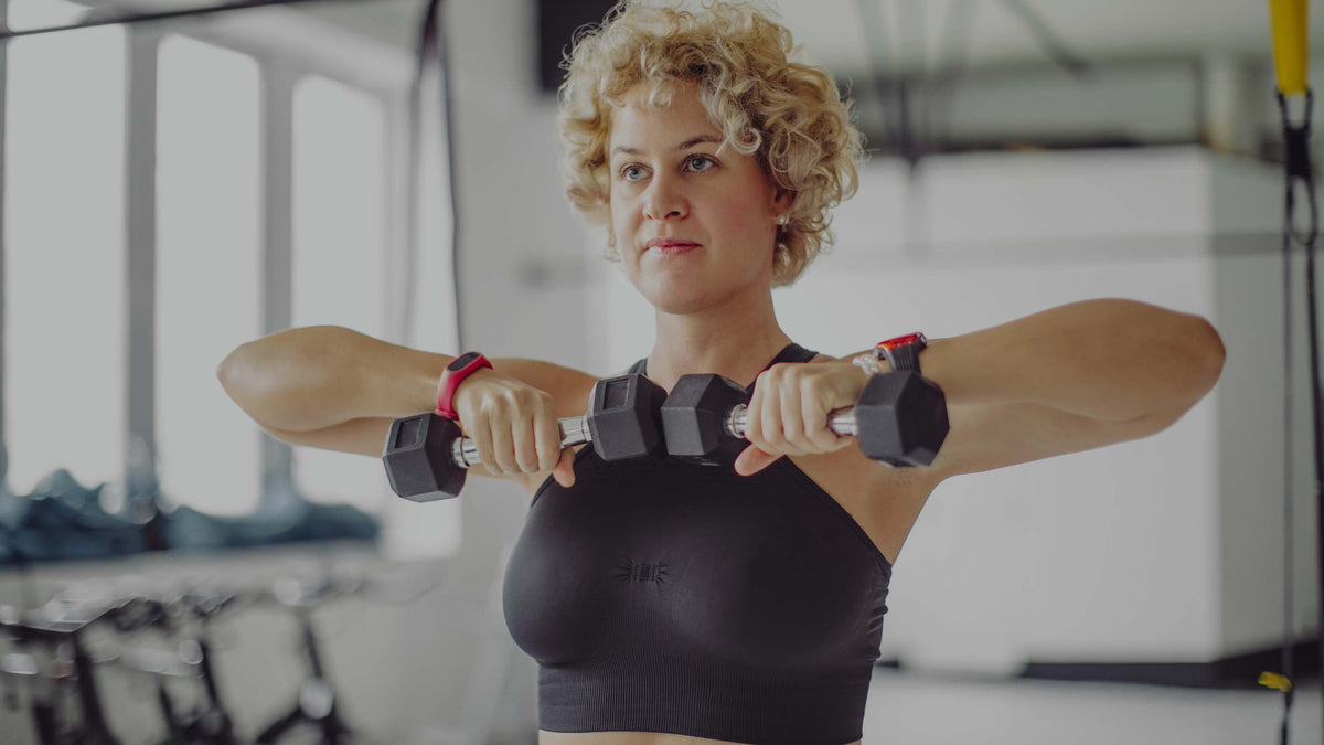 A woman with curly blonde hair in a black sports bra holds dumbbells at her shoulders in a bright gym setting.