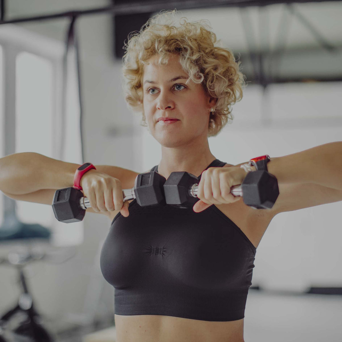 A woman with curly blonde hair wearing a black sports bra and holding dumbbells across her upper chest in a bright gym setting.