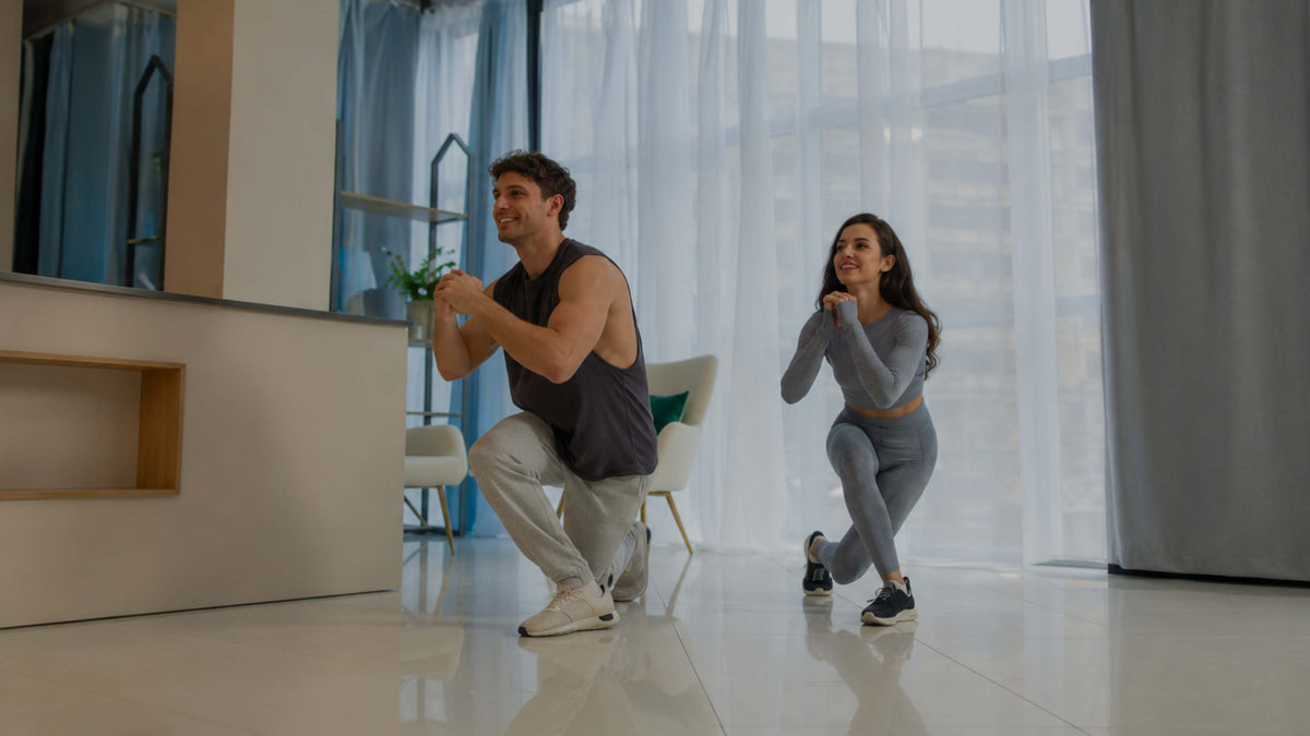 A man and a woman in athletic wear perform a squat exercise in a bright, modern indoor space with large windows and sheer curtains.