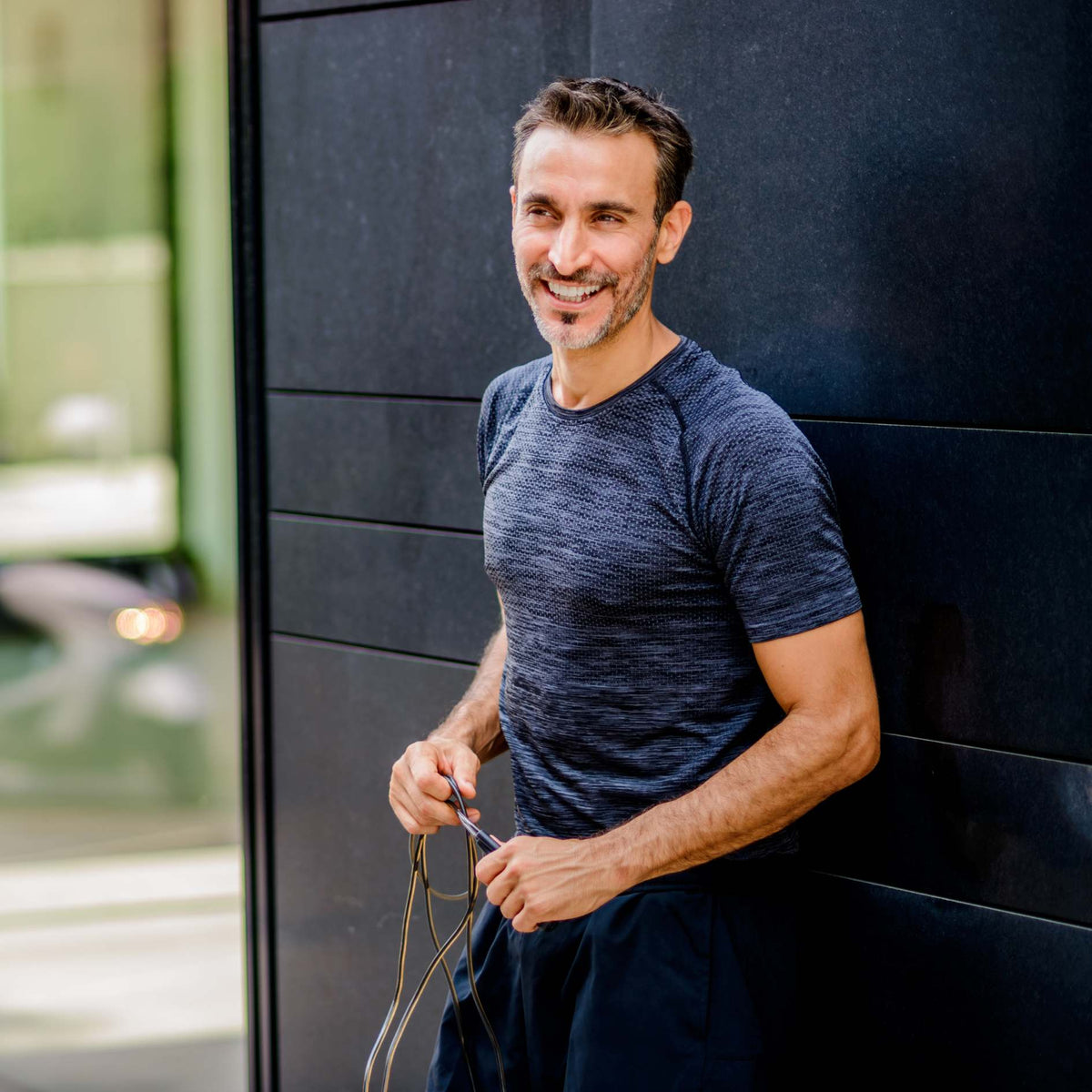 A smiling man wearing a dark heathered blue athletic t-shirt leans against a dark paneled wall, holding a pair of earbuds in his hands.