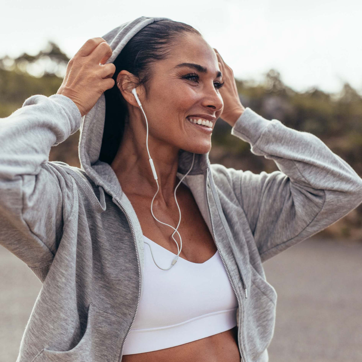 A woman outdoors wearing a light gray zip-up hoodie over a white sports bra, with white wired earphones in her ears and her hands adjusting the hood.