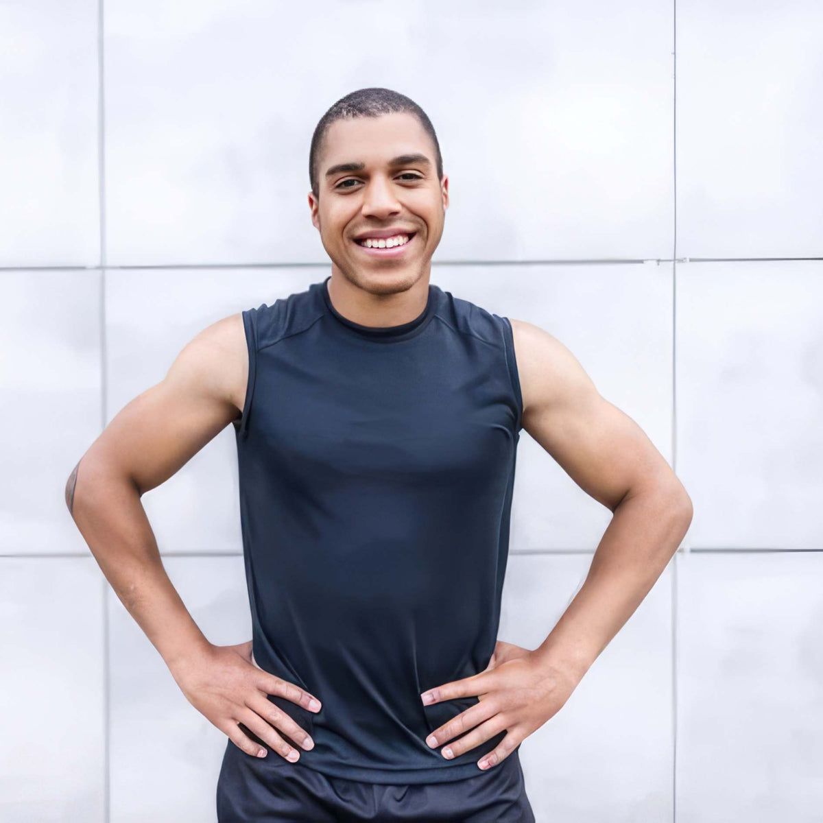 A smiling man wearing a dark navy sleeveless athletic shirt stands with hands on hips against a light gray tiled wall.