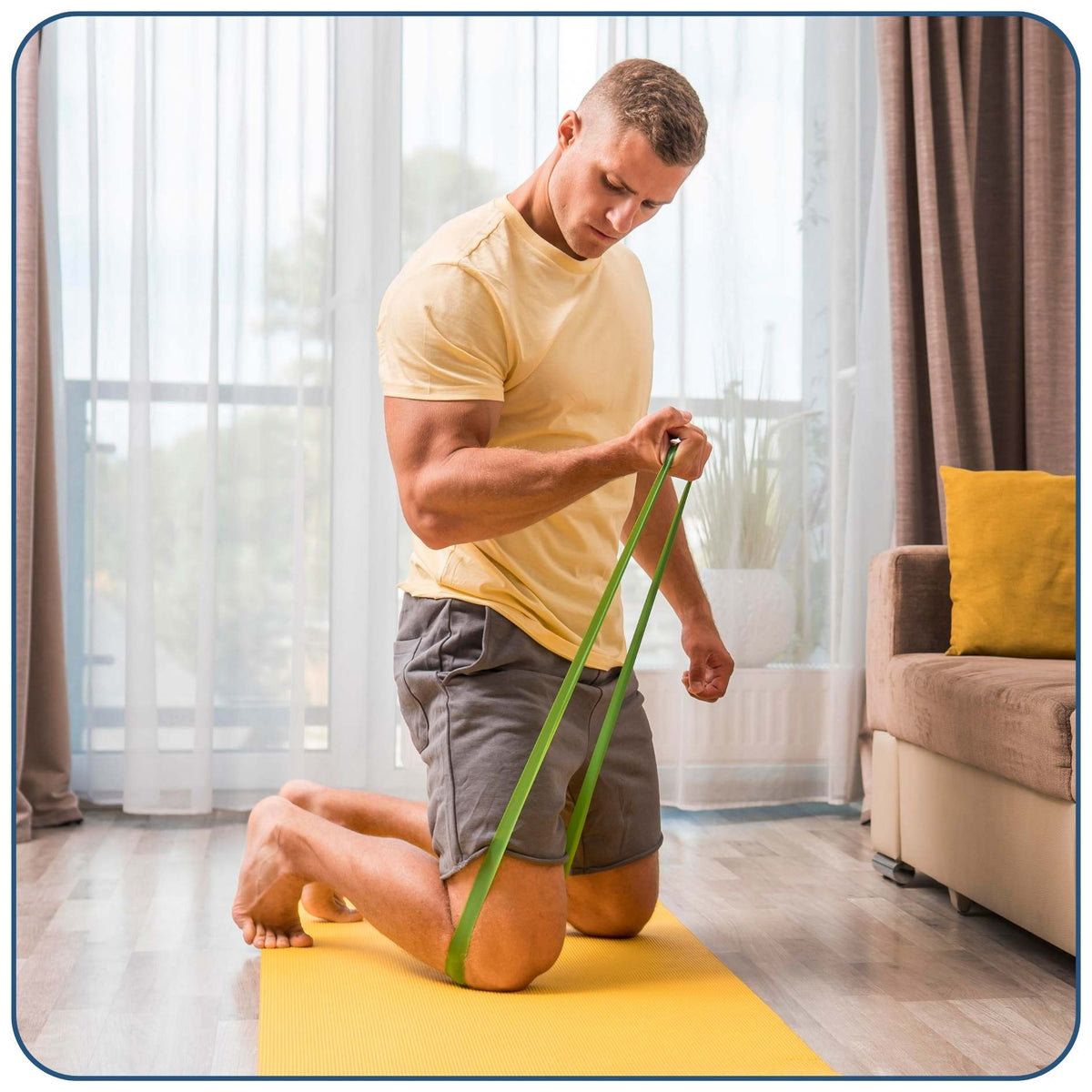 A man kneels on a yellow mat in a bright living room, using a green resistance band to work on his leg and hip. He is wearing a light yellow t-shirt and gray shorts.