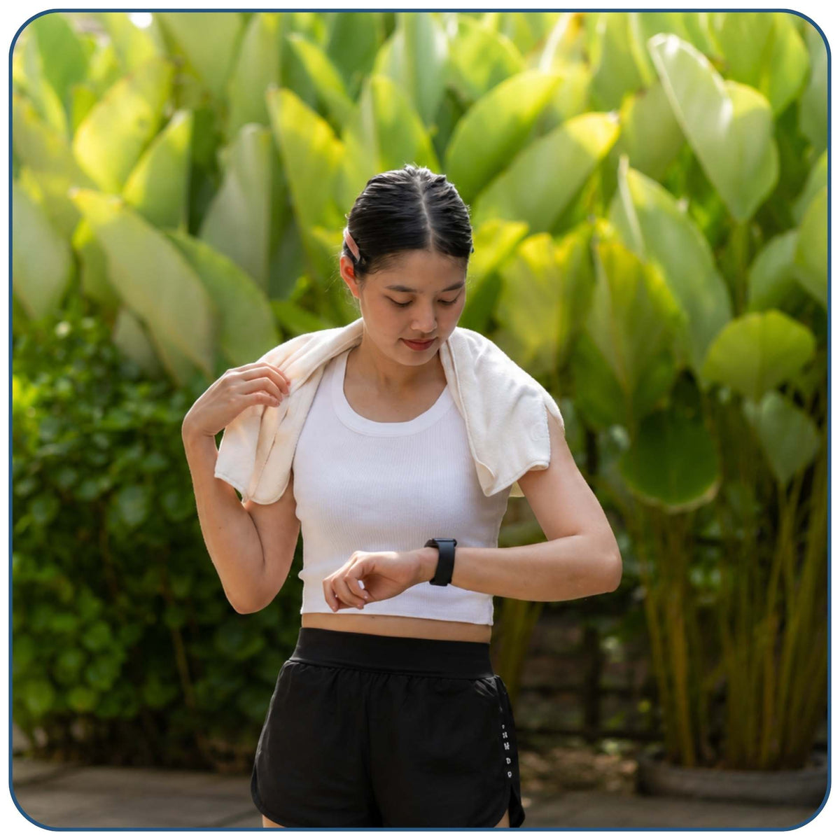 A woman outdoors in front of large green leaves, wearing a white sports tank top and black athletic shorts, with a white towel draped over her shoulders and a smartwatch on her wrist.